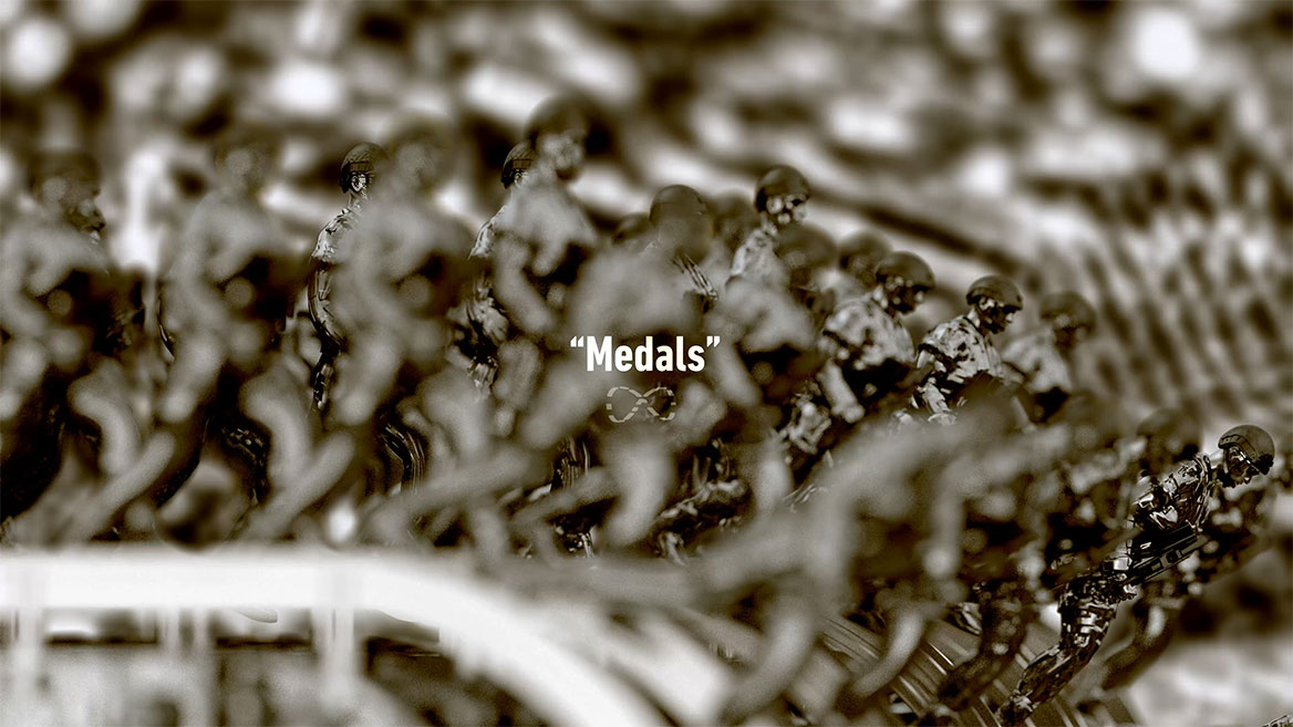 Standbild aus der Animation. Halbtotale auf die Soldatenreihen, von der Seite gesehen. Das englische Wort -Medals- steht in weißer Typografie zentral im Bild und ist mit An- und Abführungszeichen gesetzt. Darunter ist das mathematische Unendlichkeitszeichen abgebildet, dass in der Animation fast durchweg um eine unsichtbare, vertikale Achse rotiert.